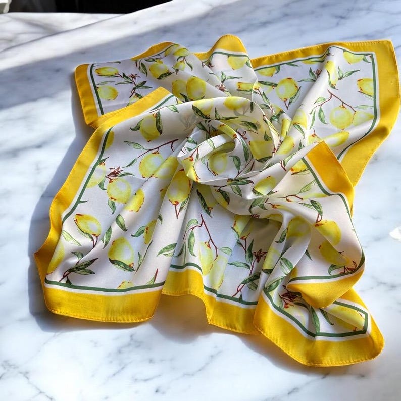 May include: A white silk scarf with a yellow border and a lemon print. The lemons are yellow with green leaves and are arranged in a repeating pattern.