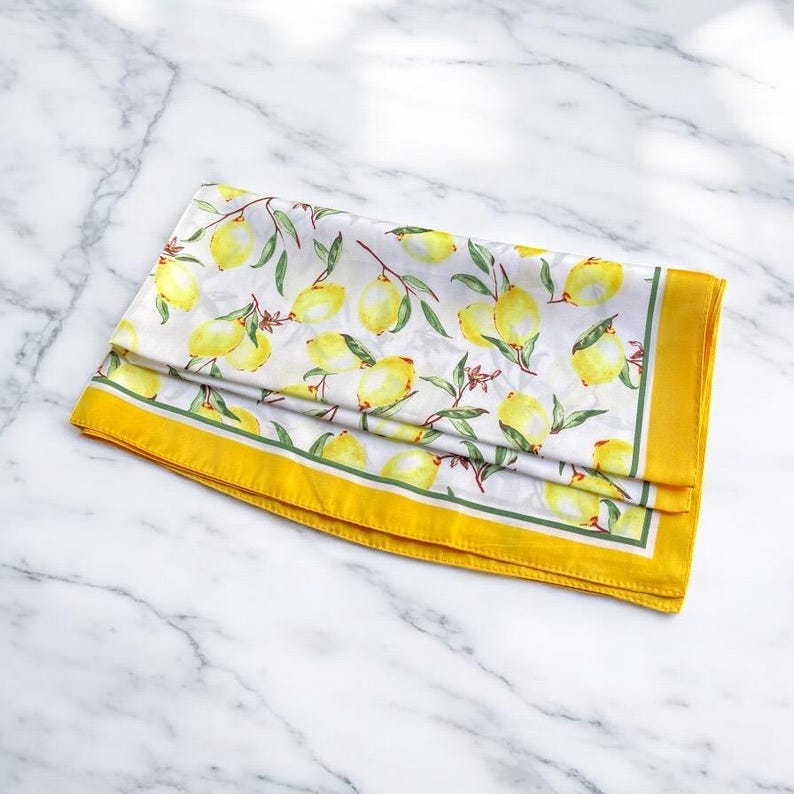 May include: A white silk scarf with a yellow lemon print and a yellow border. The scarf is folded and lying on a white marble surface.