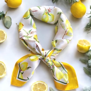 May include: A white silk scarf with a yellow border and a lemon print. The scarf is tied in a knot and the ends are hanging down. There are lemons around the scarf.