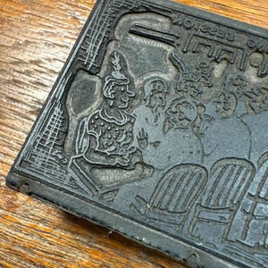 Puede incluir: Un bloque de impresi&oacute;n rectangular gris oscuro con un detallado relieve de un grupo de personas, incluyendo figuras en sillas. La imagen tiene una est&eacute;tica vintage, con texto visible y detalles intrincados.
