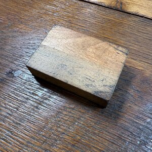 Puede incluir: Una peque&ntilde;a caja rectangular de madera con acabado natural. La caja es de madera marr&oacute;n claro con vetas m&aacute;s oscuras y grano visible. La caja est&aacute; sobre una superficie de madera.