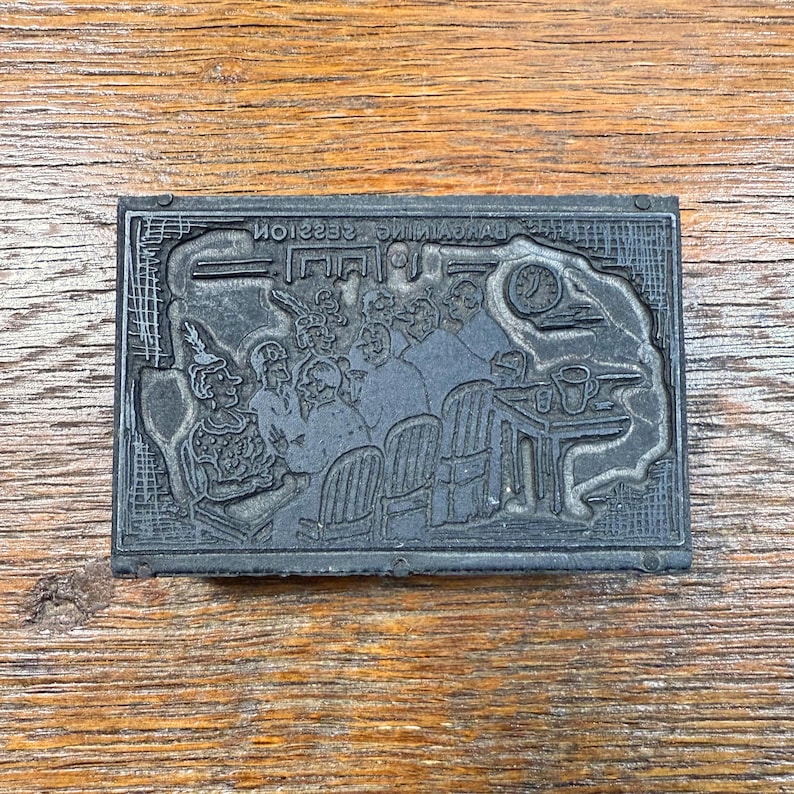 Puede incluir: Un bloque de impresi&oacute;n rectangular gris oscuro con una ilustraci&oacute;n detallada de un grupo de personas. La imagen incluye figuras sentadas en sillas y una mesa con un reloj. El texto "THE BANKING SESSION" est&aacute; impreso al rev&eacute;s en la parte superior.