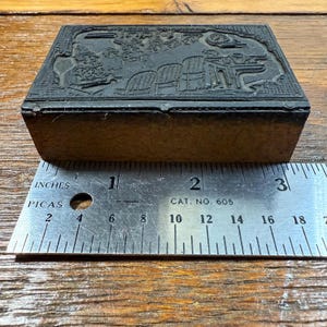 Puede incluir: Un bloque de impresi&oacute;n rectangular vintage con un dise&ntilde;o en relieve detallado. El bloque est&aacute; hecho de material oscuro y se encuentra sobre una superficie de madera. Una regla met&aacute;lica con medidas en pulgadas y picas se coloca junto al bloque. La regla dice "CAT. NO. 605".