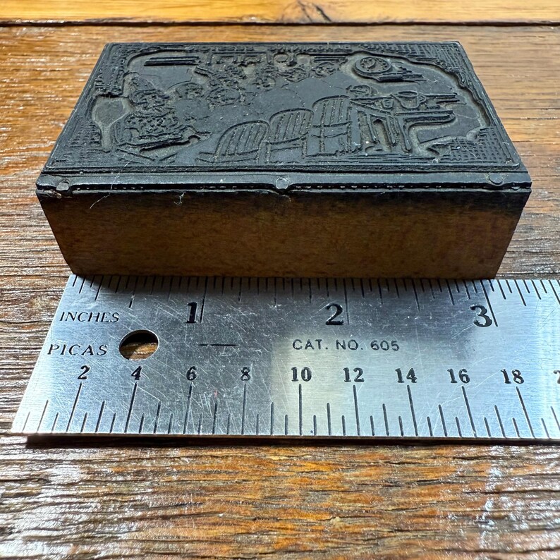 Puede incluir: Un bloque de impresi&oacute;n rectangular vintage con un dise&ntilde;o en relieve detallado. El bloque est&aacute; hecho de material oscuro y se encuentra sobre una superficie de madera. Una regla met&aacute;lica con medidas en pulgadas y picas se coloca junto al bloque. La regla dice "CAT. NO. 605".