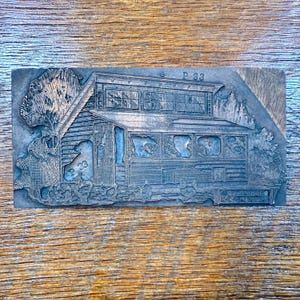 Puede incluir: Un bloque de impresión rectangular de metal gris con un relieve detallado de una casa rústica con tejado inclinado, ventanas y follaje circundante. El texto "P 33" es visible. El bloque se utiliza probablemente para la impresión tipográfica.