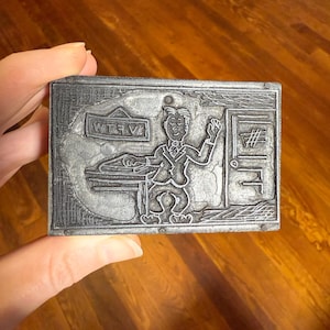Puede incluir: Un bloque de impresión rectangular de metal plateado con un hombre de dibujos animados en un escritorio con las letras "WTEV" arriba. El hombre está saludando y una puerta con un símbolo de hashtag está a la derecha.