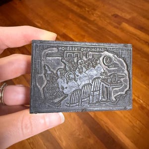 Puede incluir: Un bloque de impresi&oacute;n rectangular de metal gris con una ilustraci&oacute;n detallada de un grupo de personas. La imagen incluye figuras sentadas en mesas y un reloj. El texto "EVENING SESSION" es visible en la parte superior.