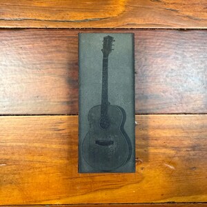 Puede incluir: Un bloque rectangular de madera gris oscuro con una ilustración detallada de una guitarra acústica. La guitarra está representada en un estilo vintage, con cuerdas, boca y clavijero visibles. El bloque descansa sobre una superficie de madera.