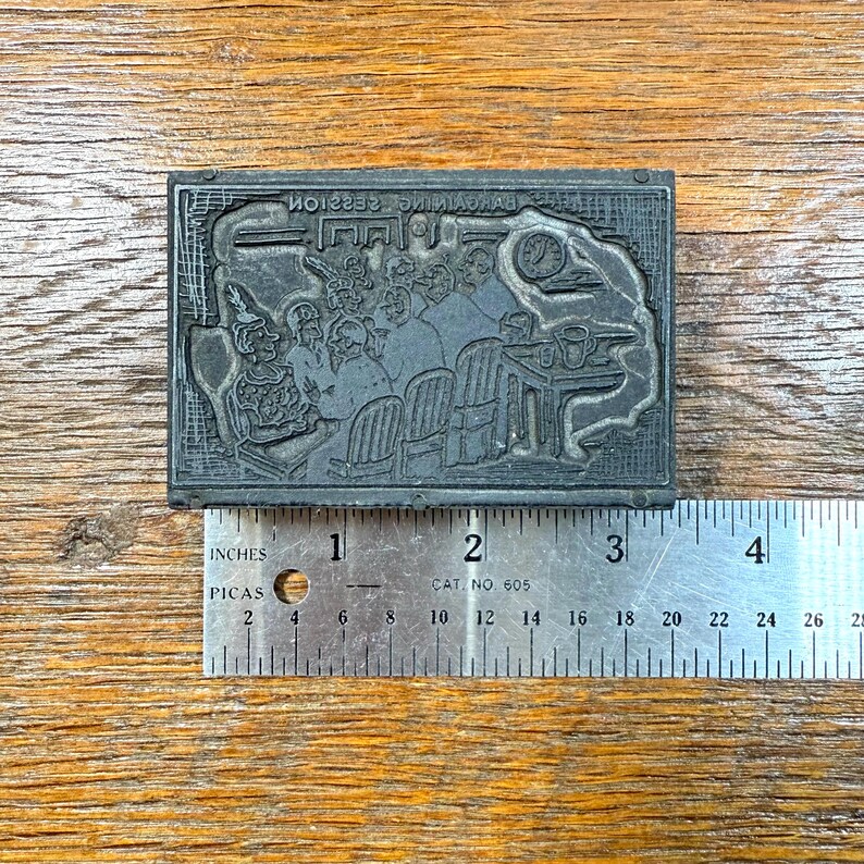 Puede incluir: Un bloque de impresi&oacute;n rectangular gris oscuro con una imagen en relieve detallada de un grupo de personas sentadas alrededor de una mesa. El bloque mide aproximadamente 10 cm de largo y tiene el texto "BACKYARD SESSION" en la parte superior.