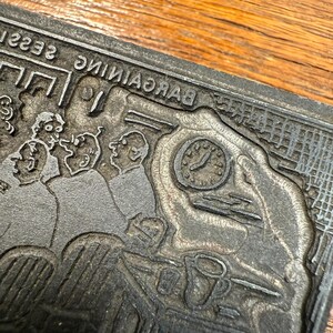 Puede incluir: Un bloque de impresi&oacute;n de metal vintage con una ilustraci&oacute;n detallada de un grupo de personas, un reloj y la palabra "BARGAINING" al rev&eacute;s. El bloque gris muestra signos de desgaste, lo que sugiere su edad y uso.