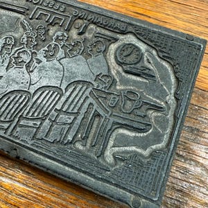Puede incluir: Un bloque de impresi&oacute;n rectangular de metal gris oscuro con una ilustraci&oacute;n en relieve detallada. La imagen representa a un grupo de personas sentadas alrededor de mesas, con un reloj y tazas de caf&eacute; visibles. El bloque est&aacute; sobre una superficie de madera.