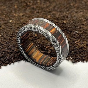 May include: A handcrafted ring with a unique design. The ring features a band of layered wood in shades of orange, brown, and black, encased in a patterned metal. The ring is set against a textured brown surface.