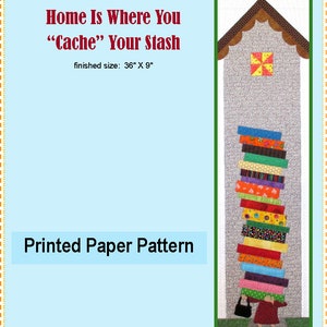 May include: A printed paper pattern for a quilt featuring a house with a stack of colorful books and a bird on the roof. The text "Home Is Where You "Cache" Your Stash" is printed above the house. The finished size of the quilt is 36 inches by 9 inches.