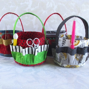 May include: Four fabric baskets with handles, each with different patterns and colors. The baskets have multiple pockets and are filled with various sewing supplies, including scissors, needles, and pens.