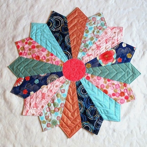 May include: A colorful quilted table runner with a starburst design. The runner features a variety of fabrics in shades of pink, blue, green, and orange. The center of the runner is a solid red circle.