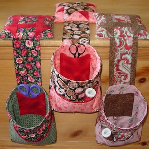 Könnte beinhalten: Vier hängende Stoffaufbewahrungstaschen mit roten, rosa, braunen und grünen Blumenmustern. Jede Tasche hat ein kleines Stoffquadrat im Inneren, und einige haben eine Garnrolle und eine Schere.