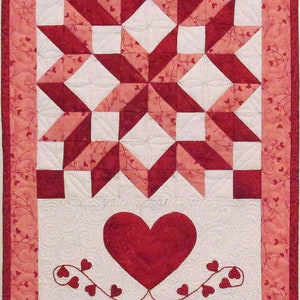 Könnte beinhalten: Eine rote und weiße Quiltdecke mit einem Sternmuster und einem roten Herzen mit Ranken am unteren Rand. Die Quiltdecke hat einen rosa Rand mit einem kleinen Herzmuster.