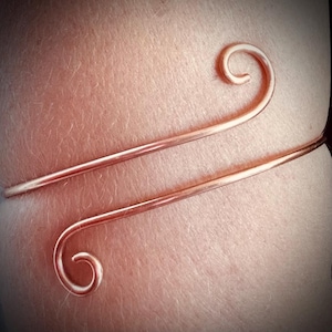 May include: A copper-colored arm cuff with a minimalist design. The cuff features two parallel, thin bands that curve around the arm, with decorative spiral ends. The jewelry is designed to be worn on the upper arm.