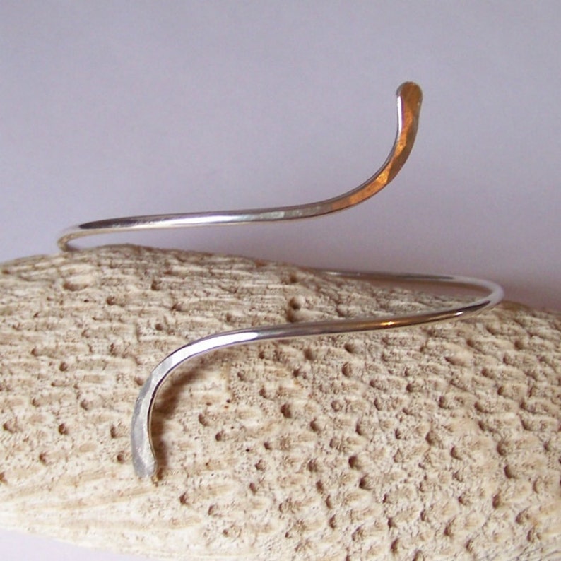 May include: A silver bangle bracelet with a simple, curved design. The bracelet is made of a single piece of wire that is twisted and bent into a spiral shape.