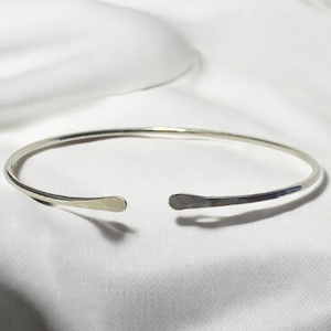May include: A silver-colored open cuff bracelet. The bracelet has a hammered texture on the ends and is set against a white background. The bracelet is a simple, elegant piece of jewelry.