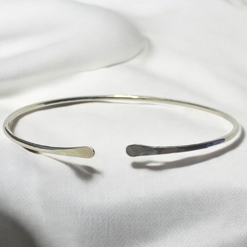 May include: A silver-colored open cuff bracelet. The bracelet has a hammered texture on the ends and is set against a white background. The bracelet is a simple, elegant piece of jewelry.