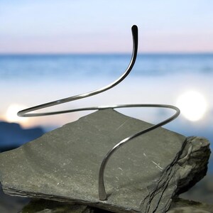 May include: A silver-toned metal arm cuff or neck piece featuring a continuous, spiraling, open-ended design. The smooth, polished wire curves upward and downward in a fluid, wave-like motion, creating a minimalist, sculptural silhouette.