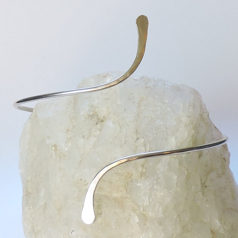 May include: A silver-colored metal arm cuff featuring a spiral design with two tapered, flattened ends. The wire is thin and smooth, curving around to create an open, adjustable shape that rests against the surface of a textured, translucent stone.