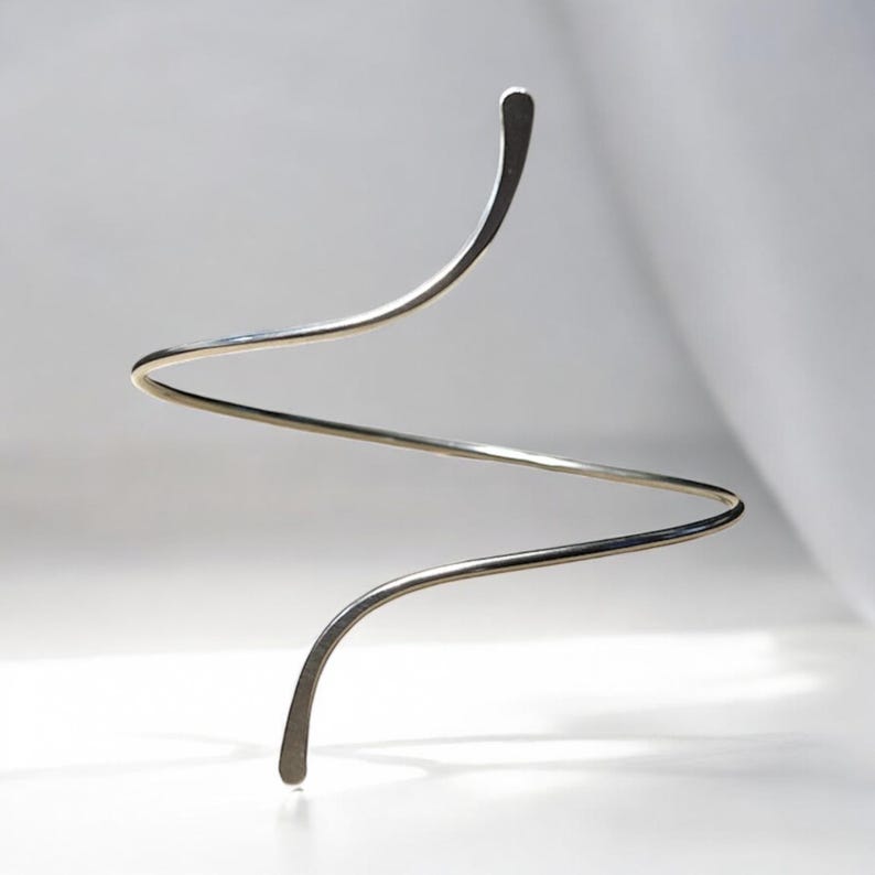 May include: A silver-toned metal arm cuff featuring a continuous, open-ended spiral design. The piece is constructed from a single, smooth, slender wire that curves into a wave-like shape, tapering slightly at both rounded ends for a clean finish.