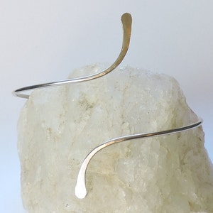 May include: A silver-colored metal arm cuff featuring a spiral design with two tapered, flattened ends. The wire is smooth and polished, curving around the form in a continuous, open-ended shape that creates a sleek, minimalist aesthetic.