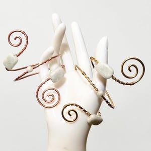 May include: Two wire-wrapped arm cuffs featuring spiral ends and light-colored rectangular stone accents. One cuff is crafted from copper-toned wire, while the other is made from gold-toned wire, both showcasing intricate coiled metal designs.