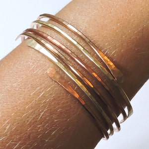 May include: A set of six hammered metal bangles in shades of gold and copper. The bangles are stacked on a person's wrist.