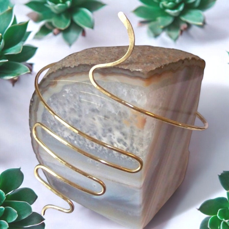 Pu&ograve; includere: Un braccialetto per braccio in metallo dorato con un design a spirale. Il braccialetto &egrave; esposto su una superficie in pietra naturale.