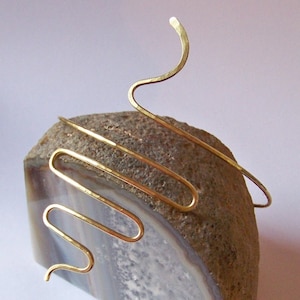 Pu&ograve; includere: Un cerchietto per capelli in metallo dorato con un design ondulato e astratto. Il cerchietto &egrave; mostrato su una pietra di agata marrone e bianca.