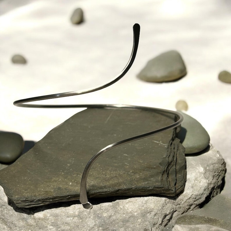 May include: A silver-toned metal neck collar featuring a continuous, open spiral design. The piece is constructed from a single, slender, smooth wire that curves upward in a fluid, circular motion, creating a minimalist and open-ended silhouette.