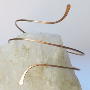 Può includere: Un semplice braccialetto in filo di rame con un design a spirale. Il braccialetto è mostrato su una roccia bianca.