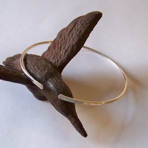 May include: A minimalist silver-toned bracelet with open ends, resting on a textured, dark brown, organic-shaped object. The bracelet's simple design and polished finish create a modern, elegant aesthetic. The bracelet is a piece of jewelry.