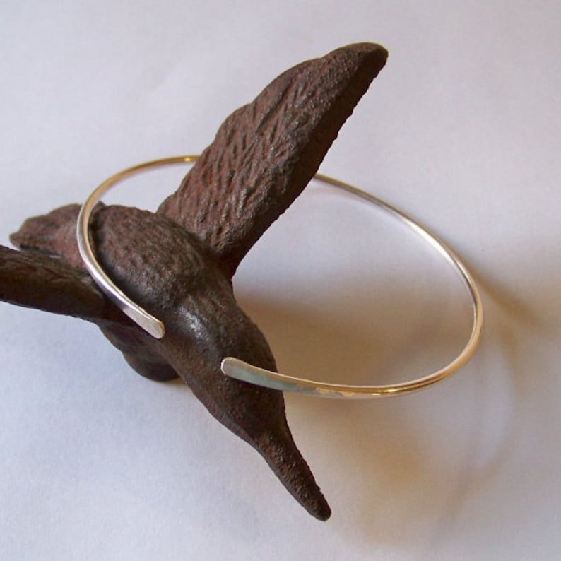 May include: A minimalist silver-toned bracelet with open ends, resting on a textured, dark brown, organic-shaped object. The bracelet's simple design and polished finish create a modern, elegant aesthetic. The bracelet is a piece of jewelry.