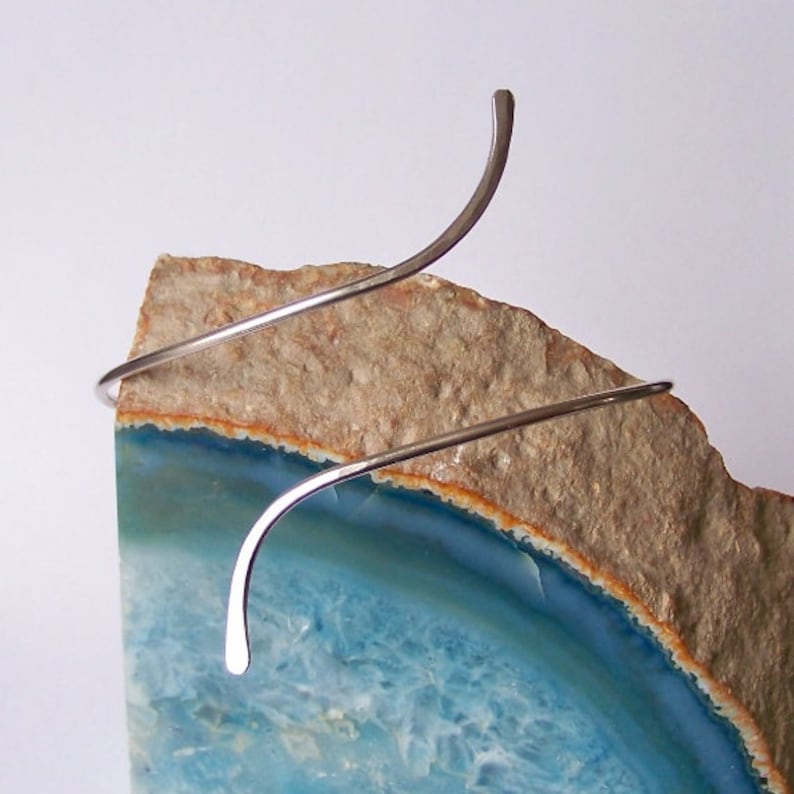 May include: A silver wire bangle bracelet with a curved design, displayed on a slice of agate with blue and brown colors.