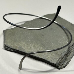 May include: A silver-toned metal choker necklace featuring a continuous, open-ended spiral design. The thin, smooth wire forms a circular shape that curves upward at one end, creating a sleek, minimalist silhouette with a polished, reflective finish.