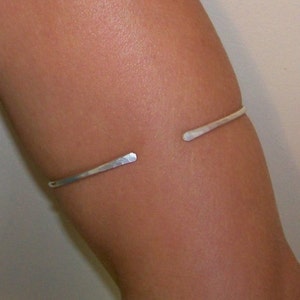 May include: A silver-colored open cuff bracelet. The bracelet is a simple, minimalist design with a hammered texture. The ends of the bracelet are slightly flattened and open, creating a modern look. The bracelet is worn on an arm.