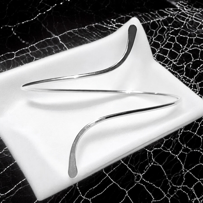 May include: A silver bangle bracelet with a unique, curved design. The bracelet is made of a single, continuous piece of metal that is twisted and shaped to create a modern and stylish look.