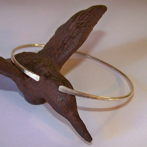 May include: A silver-colored, open cuff bracelet with hammered texture. The bracelet has a simple, minimalist design with slightly flattened ends. The bracelet is displayed on a dark brown, textured surface.