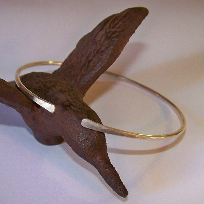 May include: A silver-colored, open cuff bracelet with hammered texture. The bracelet has a simple, minimalist design with slightly flattened ends. The bracelet is displayed on a dark brown, textured surface.