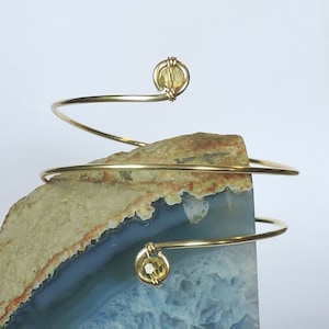 Puede incluir: Un brazalete de alambre dorado con dos cuentas de cristal amarillo. El brazalete está envuelto alrededor de una pieza de ágata.