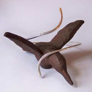 May include: A silver metal bracelet with a unique design. The bracelet is made of two pieces of silver wire that are twisted together. The bracelet is displayed on a brown, bird-shaped object.