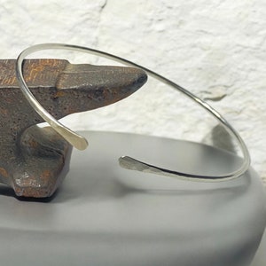 May include: A silver cuff bracelet with a hammered texture. The open-ended design features slightly flattened terminals. The bracelet is displayed on a dark gray surface, with a textured background.