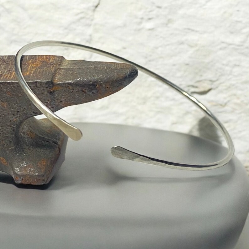 May include: A silver cuff bracelet with a hammered texture. The open-ended design features slightly flattened terminals. The bracelet is displayed on a dark gray surface, with a textured background.