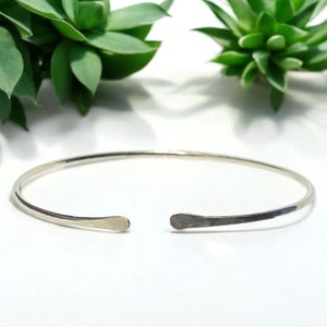 May include: A silver-colored open cuff bracelet. The bracelet has a simple, minimalist design with flattened, rounded ends. The background features green succulent plants against a white surface.