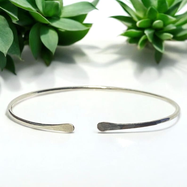 May include: A silver-colored open cuff bracelet. The bracelet has a simple, minimalist design with flattened, rounded ends. The background features green succulent plants against a white surface.
