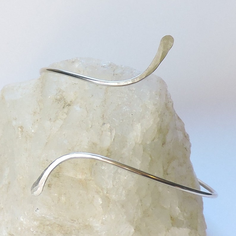 May include: A silver-colored metal arm cuff featuring a continuous, winding, wave-like design with two tapered, flattened ends. The metal surface has a smooth, reflective finish that curves around the form, creating an open, adjustable shape.